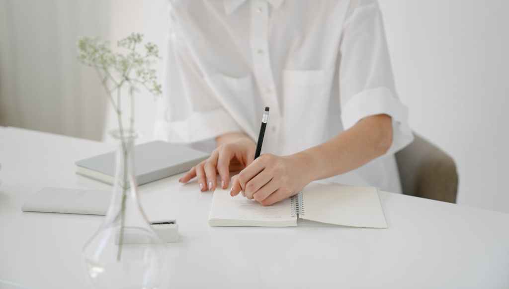 A woman is writing something on her notebook at her desk. There is a flower in a vase.
